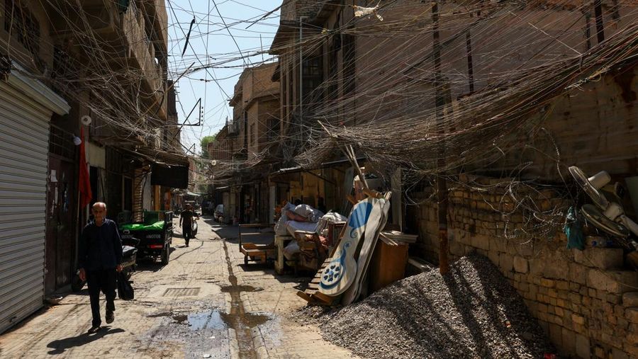 Orang-orang berjalan di sepanjang jalan tua sementara jalinan kabel listrik yang kusut dari generator yang memasok listrik ke rumah-rumah tergantung di atas gedung-gedung, di Baghdad, Irak, 10 September 2025. Orang-orang berjalan di sepanjang jalan tua sementara jalinan kabel listrik yang kusut dari generator yang memasok listrik ke rumah-rumah tergantung di atas gedung-gedung, di Baghdad, Irak, 10 September 2025. (REUTERS/Thaier Al-Sudani)