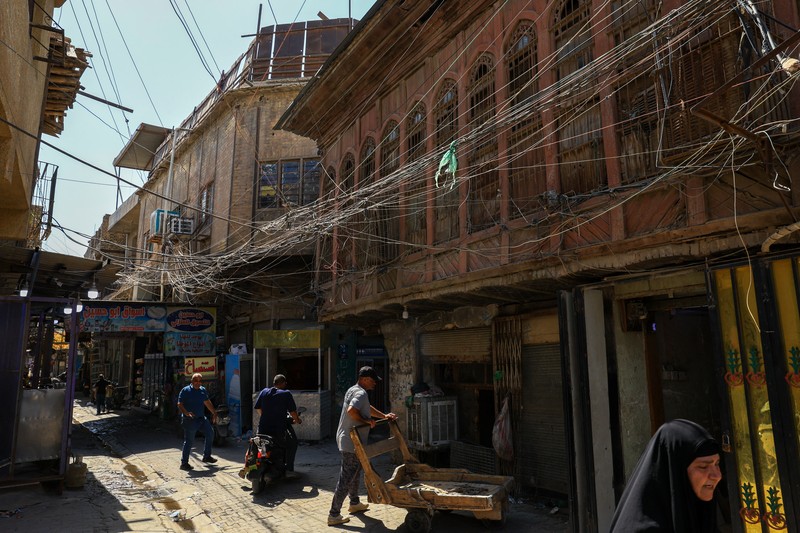 Orang-orang berjalan di sepanjang jalan tua sementara jalinan kabel listrik yang kusut dari generator yang memasok listrik ke rumah-rumah tergantung di atas gedung-gedung, di Baghdad, Irak, 10 September 2025. (REUTERS/Thaier Al-Sudani)