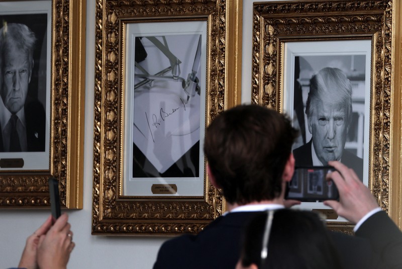 Journalists take pictures of an image of an autopen signing former U.S. President Joe Biden's signature which is displayed in place of a portrait of Biden next to portraits of U.S President Donald Trump at the 