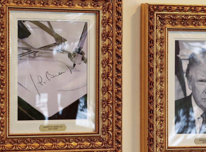 Journalists take pictures of an image of an autopen signing former U.S. President Joe Biden's signature which is displayed in place of a portrait of Biden next to portraits of U.S President Donald Trump at the 