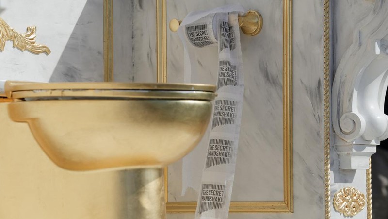 Seseorang yang mengenakan topi MAGA duduk di atas patung yang menampilkan toilet emas besar, berjudul 