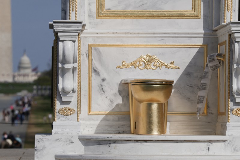 Seseorang yang mengenakan topi MAGA duduk di atas patung yang menampilkan toilet emas besar, berjudul