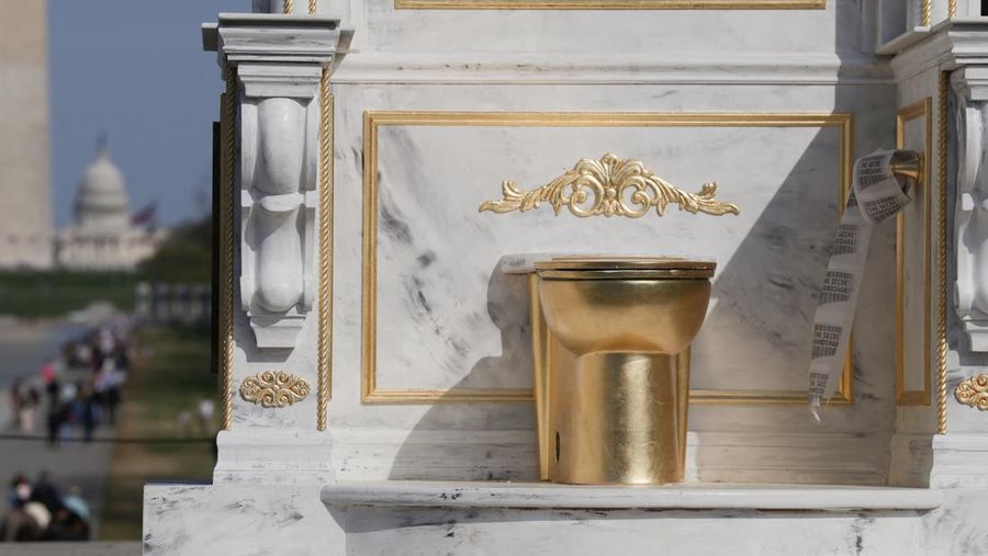 Seseorang yang mengenakan topi MAGA duduk di atas patung yang menampilkan toilet emas besar, berjudul 