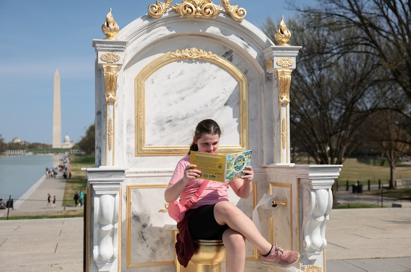 Seseorang yang mengenakan topi MAGA duduk di atas patung yang menampilkan toilet emas besar, berjudul