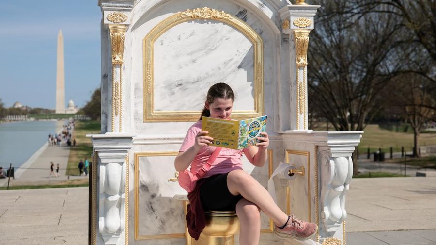 Seseorang yang mengenakan topi MAGA duduk di atas patung yang menampilkan toilet emas besar, berjudul 