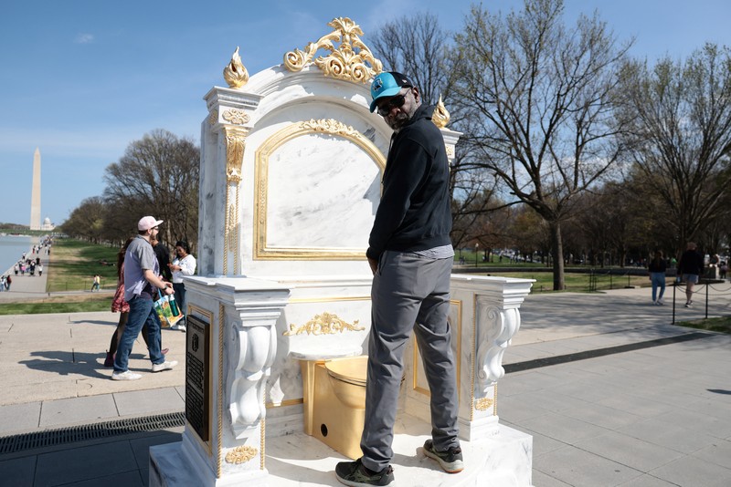 Seseorang yang mengenakan topi MAGA duduk di atas patung yang menampilkan toilet emas besar, berjudul