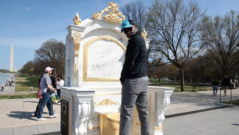 Seseorang yang mengenakan topi MAGA duduk di atas patung yang menampilkan toilet emas besar, berjudul 