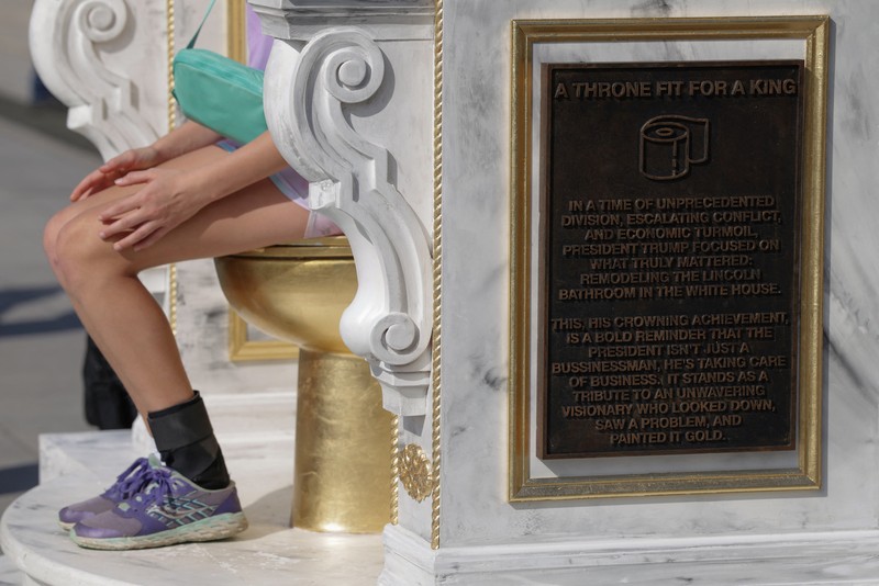 Seseorang yang mengenakan topi MAGA duduk di atas patung yang menampilkan toilet emas besar, berjudul