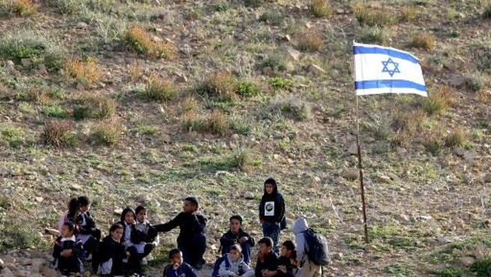 Palestinian school children stand near an Israeli flag as they returns home walking past fencing and razor wire recently erected by Israeli settlers to block the local shortcut route used to reach their school, forcing them to take a detour along the