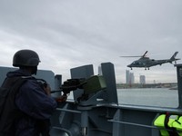 Sebuah helikopter Angkatan Laut Nigeria terbang di dekat NNS Kada, kapal operasional terbesar angkatan laut tersebut, sementara seorang anggota kru memegang senapan mesin selama latihan di luar pelabuhan yang menandai peringatan 70 tahun angkatan lau