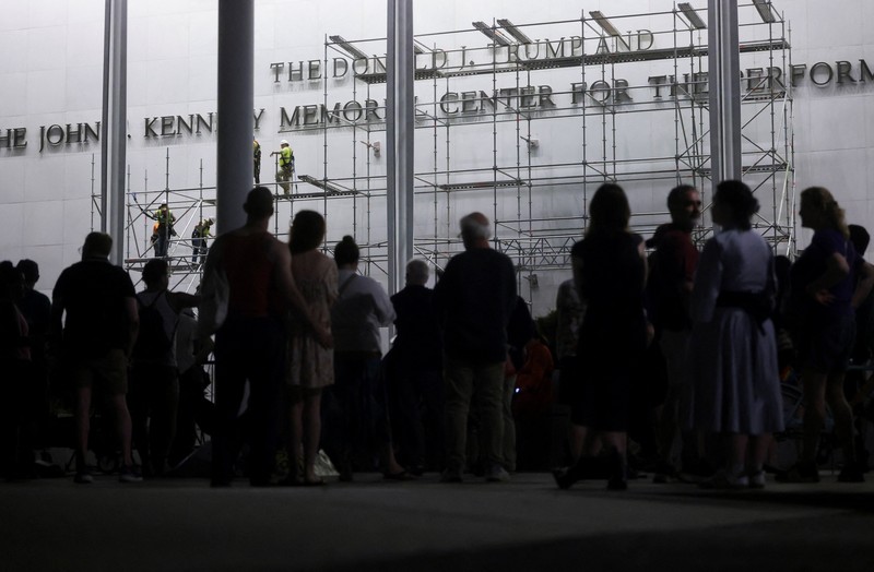 Seorang pria mengangkat bendera nasional AS sementara para pekerja menyiapkan perancah sebelum melepas tulisan dari fasad John F. Kennedy Memorial Center for the Performing Arts, menyusul perintah hakim federal untuk menghapus nama Presiden AS Donald Trump dari institusi tersebut, di Washington, D.C., AS, 12 Juni 2026. (REUTERS/Jonathan Ernst)