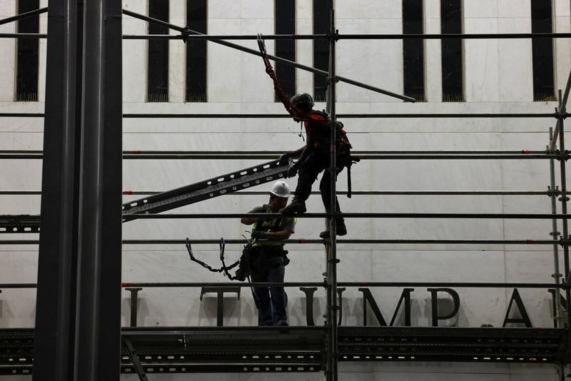 Seorang pria mengangkat bendera nasional AS sementara para pekerja menyiapkan perancah sebelum melepas tulisan dari fasad John F. Kennedy Memorial Center for the Performing Arts, menyusul perintah hakim federal untuk menghapus nama Presiden AS Donald Trump dari institusi tersebut, di Washington, D.C., AS, 12 Juni 2026. (REUTERS/Jonathan Ernst)