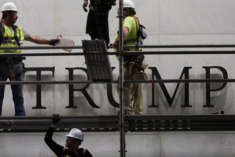 Seorang pria mengangkat bendera nasional AS sementara para pekerja menyiapkan perancah sebelum melepas tulisan dari fasad John F. Kennedy Memorial Center for the Performing Arts, menyusul perintah hakim federal untuk menghapus nama Presiden AS Donald Trump dari institusi tersebut, di Washington, D.C., AS, 12 Juni 2026. (REUTERS/Jonathan Ernst)
