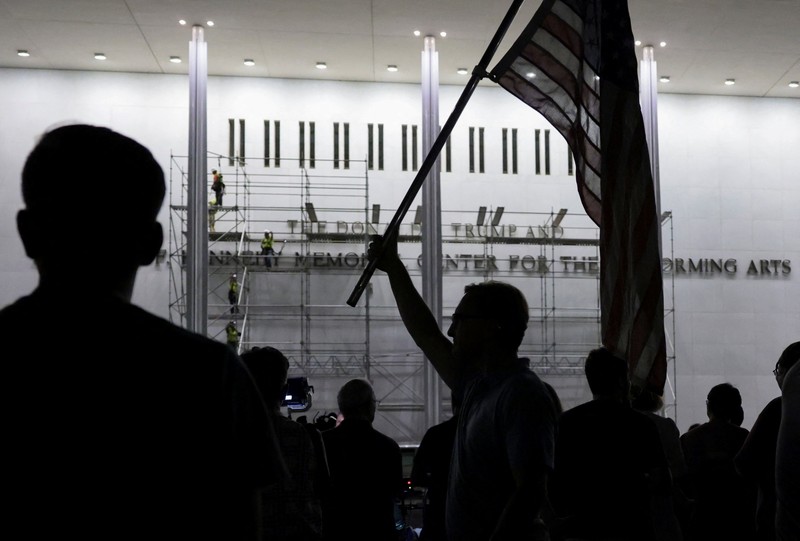Seorang pria mengangkat bendera nasional AS sementara para pekerja menyiapkan perancah sebelum melepas tulisan dari fasad John F. Kennedy Memorial Center for the Performing Arts, menyusul perintah hakim federal untuk menghapus nama Presiden AS Donald Trump dari institusi tersebut, di Washington, D.C., AS, 12 Juni 2026. (REUTERS/Jonathan Ernst)