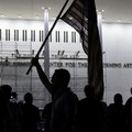 Seorang pria mengangkat bendera nasional AS sementara para pekerja menyiapkan perancah sebelum melepas tulisan dari fasad John F. Kennedy Memorial Center for the Performing Arts, menyusul perintah hakim federal untuk menghapus nama Presiden AS Donald