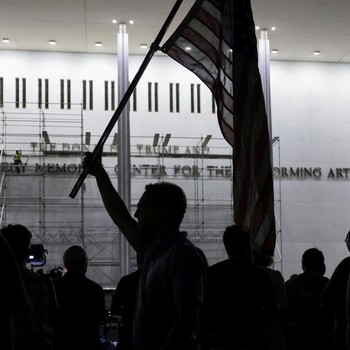 Seorang pria mengangkat bendera nasional AS sementara para pekerja menyiapkan perancah sebelum melepas tulisan dari fasad John F. Kennedy Memorial Center for the Performing Arts, menyusul perintah hakim federal untuk menghapus nama Presiden AS Donald