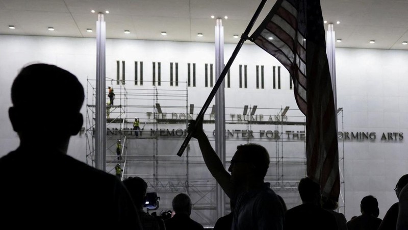 Seorang pria mengangkat bendera nasional AS sementara para pekerja menyiapkan perancah sebelum melepas tulisan dari fasad John F. Kennedy Memorial Center for the Performing Arts, menyusul perintah hakim federal untuk menghapus nama Presiden AS Donald
