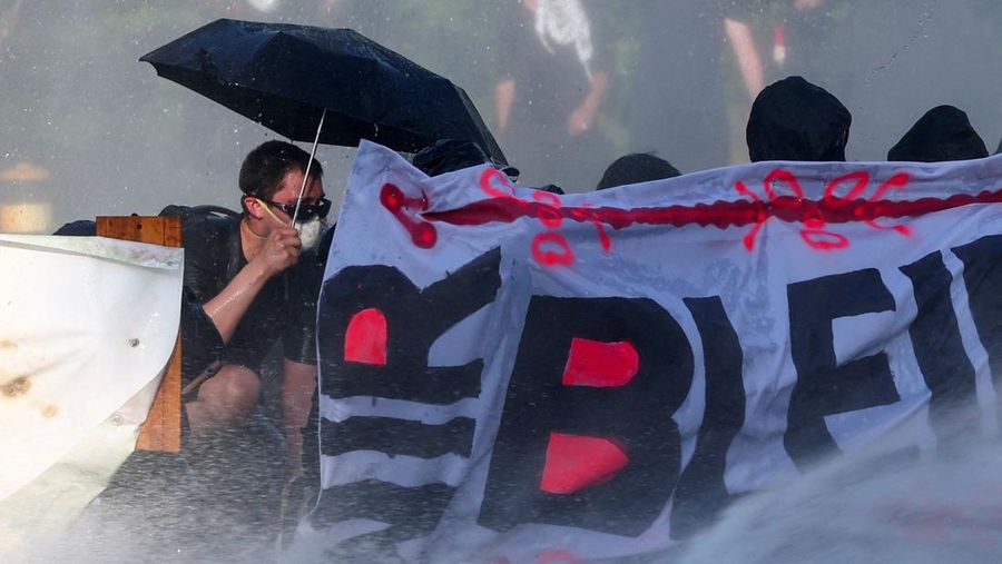 Seorang demonstran memegang payung sambil berlindung di balik spanduk saat polisi anti huru hara menyiram mereka dengan air dari meriam air selama protes menentang KTT G7 yang akan datang di Evian-les-Bains, Prancis, di Jenewa, Swiss, 14 Juni 2026.