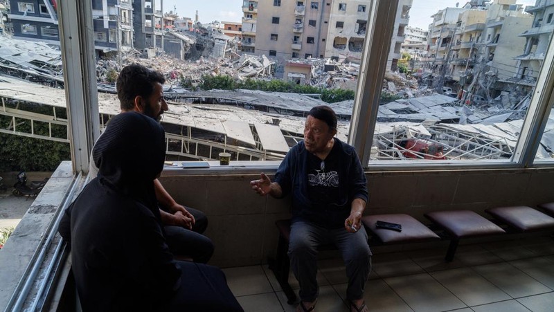 Youssef Fares, 90, whose home of more than 70 years was damaged by an Israeli airstrike, chats with fellow displaced people as they sit beside broken windows of Jabal Amel hospital, where he has taken shelter with his family, overlooking buildings da