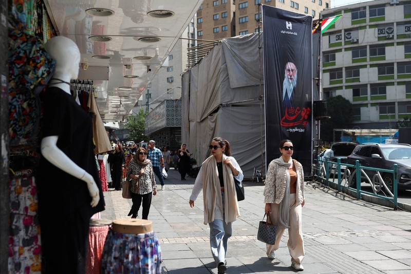Perempuan Iran berjalan melewati mural anti-AS di sebuah jalan di Teheran, Iran, Senin (15/6/2026). Meski hidup dalam ketidakpastian, sebagian warga Iran berusaha menjalani hari seperti biasa. (via REUTERS/Majid Asgaripour)