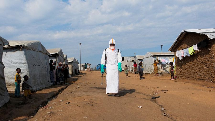 Para pengungsi menyaksikan seorang petugas kesehatan dengan perlengkapan pelindung diri (PPE) lengkap bersiap untuk mendisinfeksi area tersebut selama pemakaman korban yang diduga terinfeksi Ebola di kamp pengungsi Kigonze di Bunia, bagian timur Repu