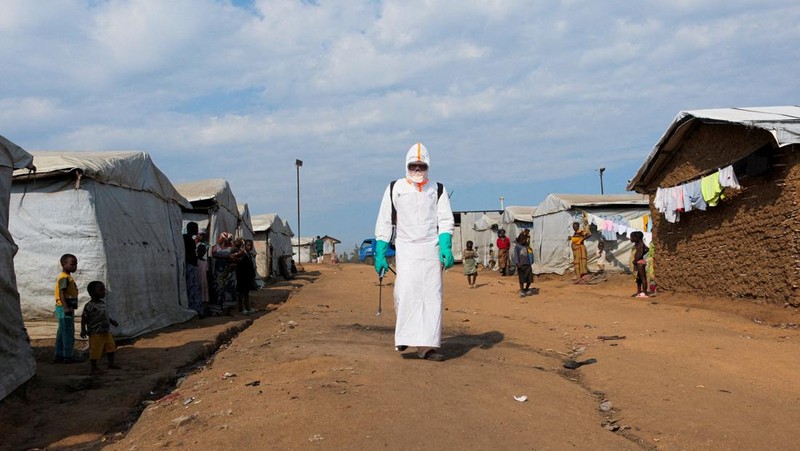 Para pengungsi menyaksikan seorang petugas kesehatan dengan perlengkapan pelindung diri (PPE) lengkap bersiap untuk mendisinfeksi area tersebut selama pemakaman korban yang diduga terinfeksi Ebola di kamp pengungsi Kigonze di Bunia, bagian timur Repu