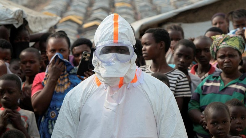 Seorang petugas kesehatan dengan alat pelindung diri (APD) berdiri di dekat para pengungsi yang menunggu pemakaman korban yang diduga terinfeksi Ebola di kamp pengungsi Kigonze, satu bulan setelah wabah dinyatakan, di Bunia, bagian timur Republik Demokratik Kongo, 18 Juni 2026.
