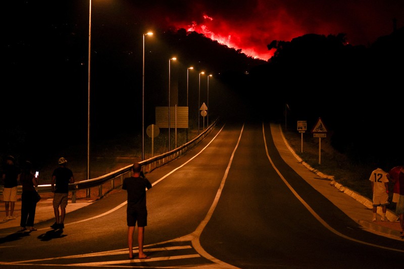 People look on as flames and smoke from a wildfire rise near Calonge, Spain, July 4, 2026. REUTERS/Bruna Casas     TPX IMAGES OF THE DAY