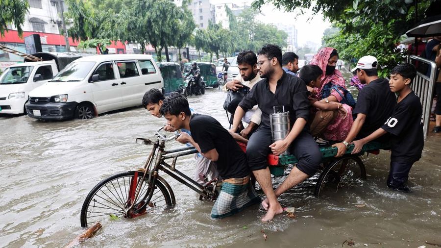 Orang-orang menyeberangi jalan yang tergenang banjir menggunakan becak setelah hujan deras di Dhaka, Bangladesh, 12 Juli 2026.
