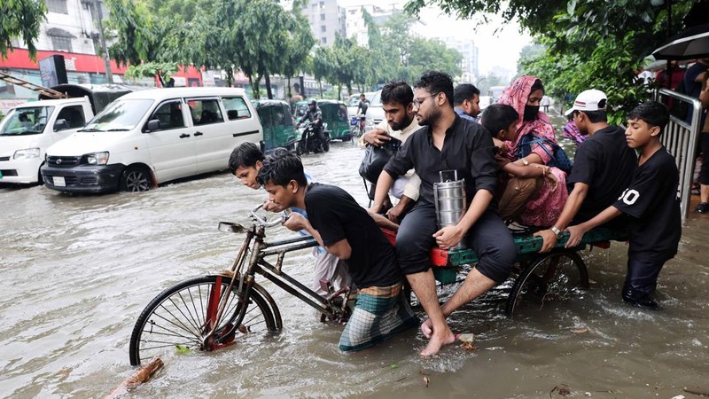 Orang-orang menyeberangi jalan yang tergenang banjir menggunakan becak setelah hujan deras di Dhaka, Bangladesh, 12 Juli 2026.