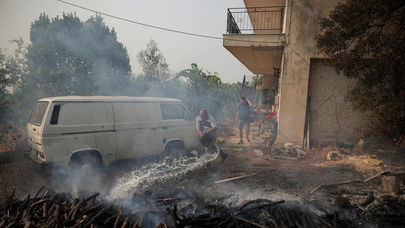 Seorang warga menyiramkan air ke kayu yang masih berasap di luar sebuah rumah setelah kebakaran hutan Boeotia melanda Veniza, Yunani, 2 Agustus 2026.