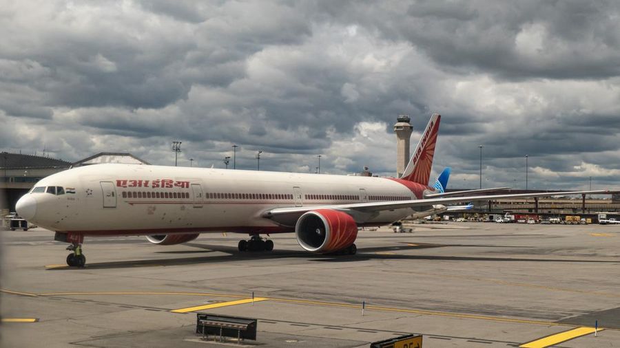 Pesawat penumpang Boeing 777 milik Air India terlihat di landasan Bandara Internasional Newark Liberty, Amerika Serikat, dengan latar belakang menara kendali dan terminal bandara di bawah langit mendung dan cuaca berawan gelap.