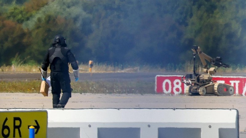 Seorang petugas polisi yang mengenakan pakaian pelindung berjalan di samping robot penjinak bahan peledak (EOD) di Bandara Leipzig/Halle di Schkeuditz, Jerman, pada 5 Agustus 2026. Langkah ini diambil setelah polisi menyatakan bahwa ahli bahan peledak telah memeriksa sebuah pesawat nirawak (drone) yang ditemukan di dekat landasan pacu, serta menyusul insiden pesawat kargo DHL yang menabrak objek tak dikenal saat sedang menanjak meninggalkan bandara pada malam sebelumnya, yang memicu pendaratan tak terjadwal di Hanover, menurut sumber yang mengetahui peristiwa tersebut.