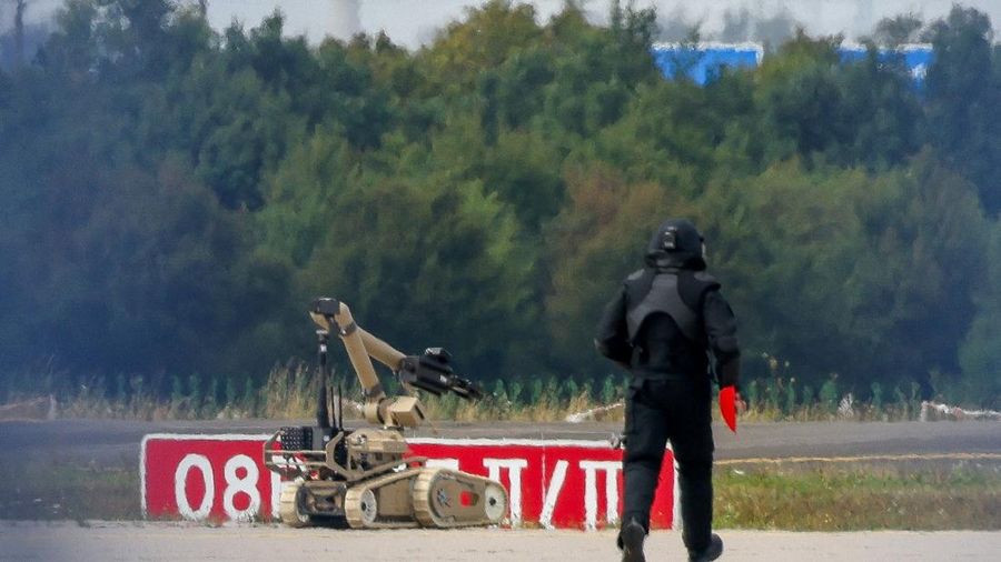 Seorang petugas polisi yang mengenakan pakaian pelindung berjalan di samping robot penjinak bahan peledak (EOD) di Bandara Leipzig/Halle di Schkeuditz, Jerman, pada 5 Agustus 2026. Langkah ini diambil setelah polisi menyatakan bahwa ahli bahan peledak telah memeriksa sebuah pesawat nirawak (drone) yang ditemukan di dekat landasan pacu, serta menyusul insiden pesawat kargo DHL yang menabrak objek tak dikenal saat sedang menanjak meninggalkan bandara pada malam sebelumnya, yang memicu pendaratan tak terjadwal di Hanover, menurut sumber yang mengetahui peristiwa tersebut.