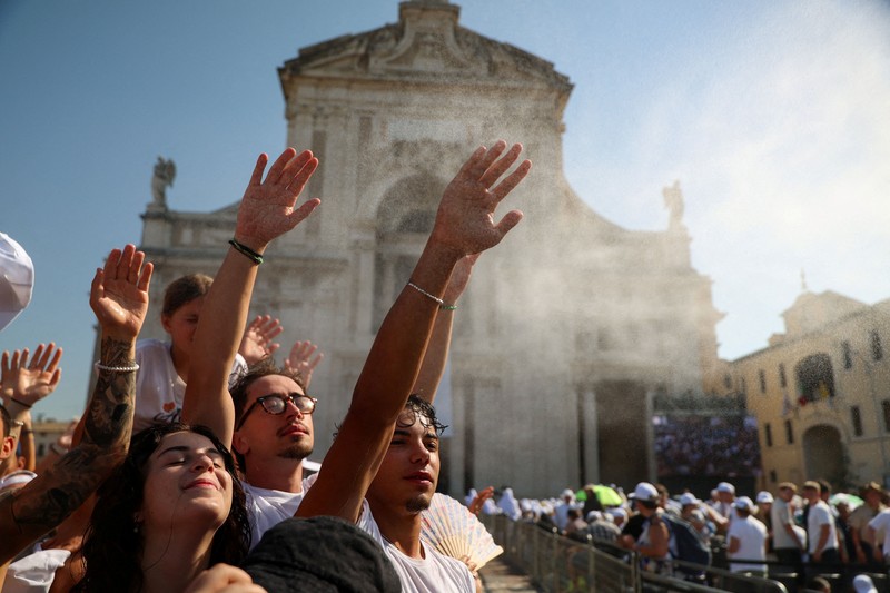 Warga mengangkat tangan sembari menyejukkan diri di tengah semburan kabut air di luar Basilika Santa Maria degli Angeli saat berkumpul untuk menyambut kunjungan Paus Leo dalam rangka peringatan 800 tahun wafatnya Santo Fransiskus, di tengah gelombang panas di Assisi, Italia, pada 6 Agustus 2026. (REUTERS/Antonio Denti)