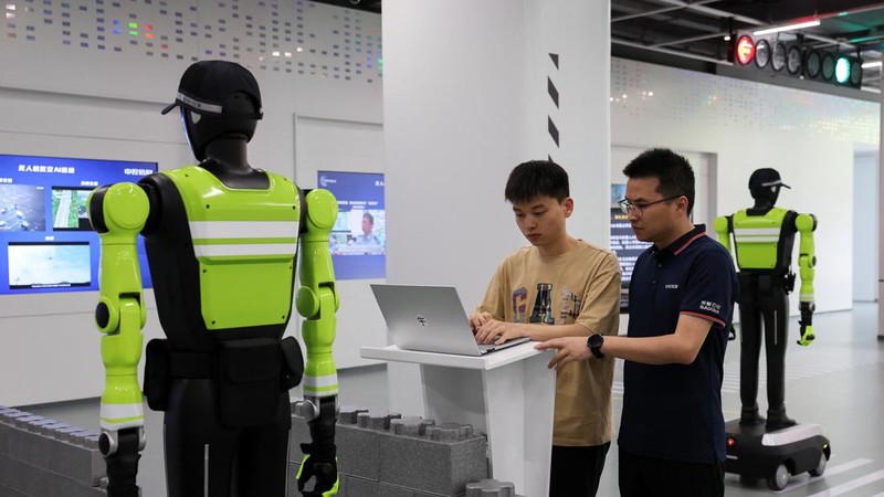 Cao Hui, manajer umum Pusat Inovasi Sistem Nirawak Cerdas SUPCON, dan seorang insinyur sedang bekerja di depan komputer sementara robot humanoid polisi lalu lintas berdiri di ruang pamer perusahaan tersebut di Hangzhou, Provinsi Zhejiang, Tiongkok, pada 21 Juli 2026.