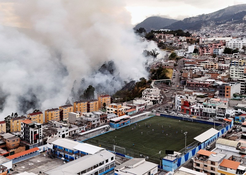 Kebakaran hutan di dekat kawasan permukiman, sementara orang-orang bermain sepak bola di sebuah lapangan di Quito, Ekuador, Minggu (13/9/2026). (REUTERS/Karen Toro)