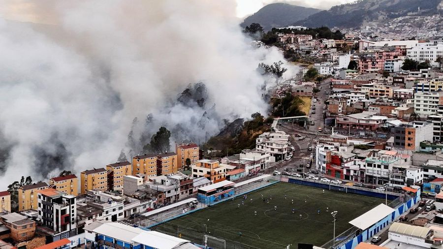 Kebakaran hutan di dekat kawasan permukiman, sementara orang-orang bermain sepak bola di sebuah lapangan di Quito, Ekuador, Minggu (13/9/2026). (REUTERS/Karen Toro)