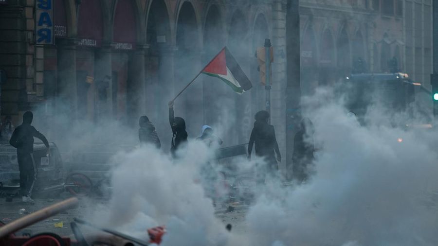 Seorang pengunjuk rasa mengibarkan bendera Palestina di tengah kepulan asap saat terjadi bentrokan dengan polisi, menyusul aksi protes menentang kenaikan biaya hidup di Bologna, Italia, pada 17 Oktober 2026.