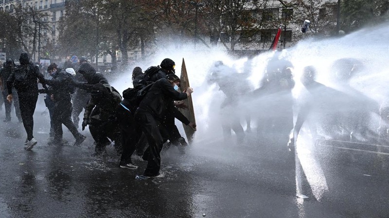 Protesters take cover behind a plywood board as French riot police use a water canon amid clashes during an authorised demonstration organised by labour unions in Paris as part of nationwide protests called by the "Yellow Vests" movement joined by Fr