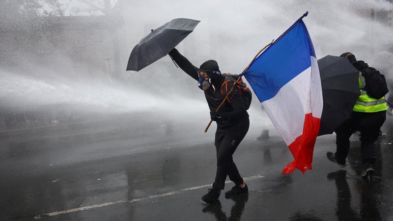 A masked prostester holding an umbrella and a French flag reacts as French police use a water canon  amid clashes during an authorised demonstration organised by labour unions in Paris as part of nationwide protests called by the "Yellow Vests" movem