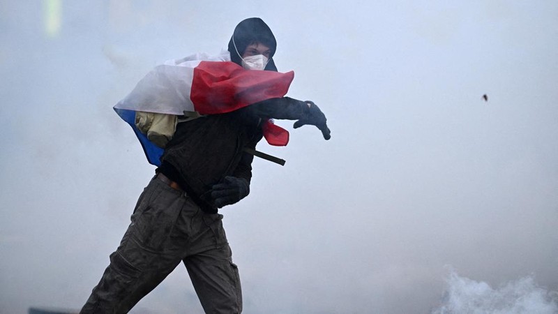 A masked protester wearing a French flag throws a tear gas submunition amid clashes with French police during an authorised demonstration organised by labour unions in Paris as part of nationwide protests called by the