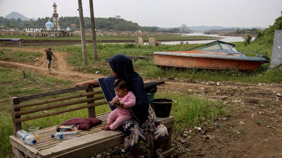 Martini, 45 tahun, menggendong keponakannya yang berusia 6 bulan. Mereka duduk di dekat sebuah perahu yang terdampar di lumpur setelah permukaan air di waduk Karian—yang memasok air untuk wilayah sekitarnya, termasuk Jakarta—menurun akibat kekeringan berkepanjangan yang dipicu oleh fenomena El Niño. Kondisi ini menyebabkan sisa-sisa rumah dan masjid di desa Somang muncul kembali ke permukaan, di Lebak, provinsi Banten, Indonesia, 23 Oktober 2026.